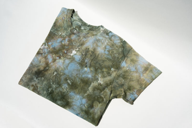 The UNA Ice Dyed Organic Short Sleeve Shirt by Broc Cellars is laid flat on a light surface, featuring a blend of muted green, blue, and brown hues. A small logo or text by Marta Elise Johansen subtly adorns the upper center, adding intrigue to its design.