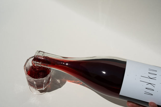 A person pours red liquid from a 2023 KouKou Cabernet Franc bottle by Broc Cellars into a glass on a light surface. The minimalist label, partly visible, suggests its made with organic grapes using carbonic fermentation.