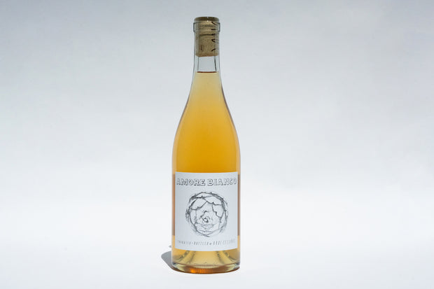 A bottle of Amore Trio wine from Broc Cellars is centered against a plain background. The label, capturing the essence of Italian table wines, boasts a minimalist design with the name and an artichoke illustration. The wine is light amber in color.