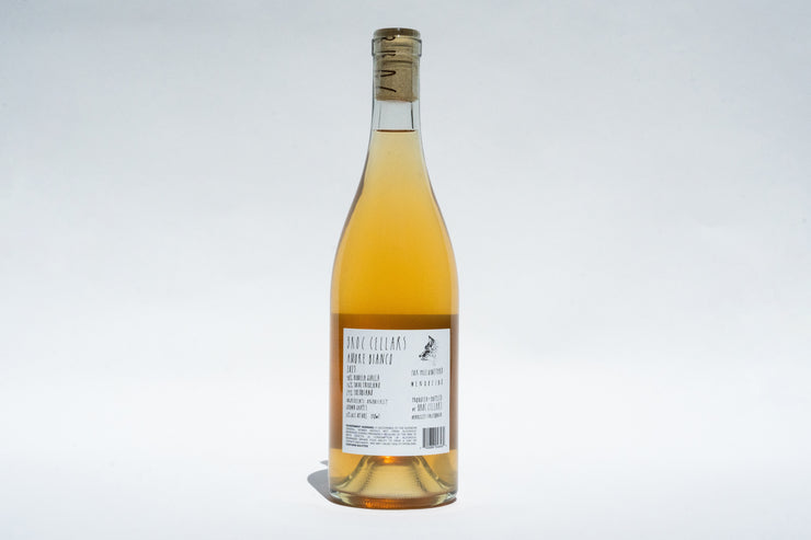 A bottle of 2023 Amore Bianco from Broc Cellars showcases a clear glass body filled with amber liquid. The cork-sealed top and rectangular white label pop against the plain background, emphasizing this exquisite Italian skin contact wine.