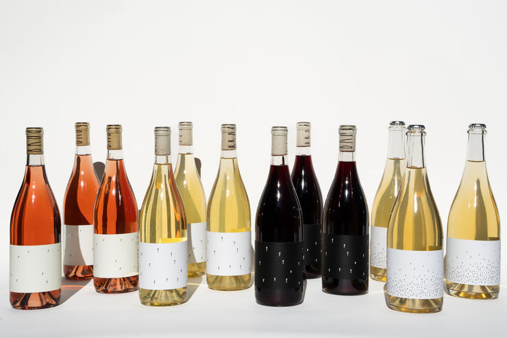Twelve bottles from Broc Cellars’ Love Case—rosé, white, and red—are arranged in two rows on a white background with blank labels and cork stoppers. Love wine? Enjoy free shipping on this exclusive set.