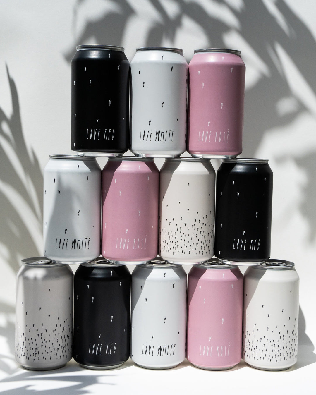 A pyramid of twelve Broc Cellars Love Can Variety Pack cans with black, white, and pink minimalist designs sits on a white surface with shadows. Arranged in three rows, they make stylish picnic drinks.