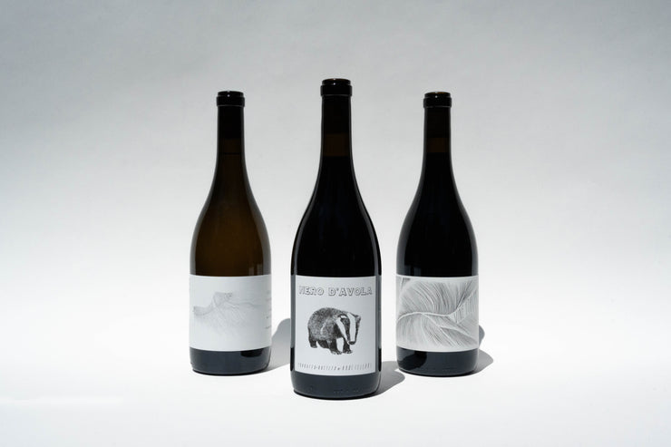 Three wine bottles, part of Chris Picks by Broc Cellars, are set against a plain background. The center bottle, labeled NERO DAVOLA, features a charming badger illustration, while the flanking bottles have minimalist leaf designs; one is a rich California Zinfandel.