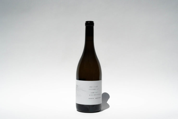 A tall, dark glass wine bottle with a minimalistic white label of the 2023 Michael Mara Chardonnay from Broc Cellars, featuring Sonoma Coast AVA, is centered against a plain background. The label has small, partially visible text and an embossed design, with the bottle casting a shadow to the right.