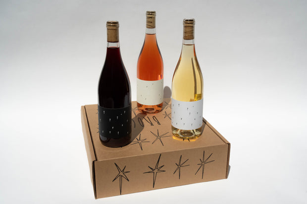 Three Broc Cellars wine bottles elegantly rest on a cardboard box, presenting the delightful Love Trio. Featuring minimalist labels with small black lines, these bottles contain dark red, Love Rosé, and white wine. Star-like designs embellish the box, adding whimsy to the display.