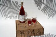 A bottle of Rosato from Broc Cellars sits invitingly with two filled wine glasses on a star-adorned gift box, while fern leaf shadows dance on the white backdrop, enhancing this elegant scene.