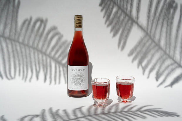 A bottle of Rosato with Wine Glasses by Broc Cellars rests on a surface with two filled glasses. Fern leaf shadows create a decorative background, complementing the rosé wine, ideal for an elegant gift box presentation.