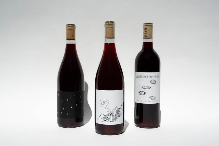 Three bottles of Broc Cellars Red Trio, featuring artistic abstract labels, stand in a row on a white surface with their corks intact and shadows cast behind. Perfect for those seeking versatile wines that inspire creativity without breaking the bank.