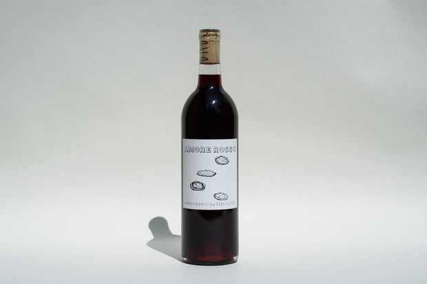 A bottle of 2023 Amore Rosso by Broc Cellars stands upright against a light background. The label showcases Amore Rosso with three oyster shell illustrations, indicating its Barbera Dolcetto blend. It is capped with a cork wrapped in foil.