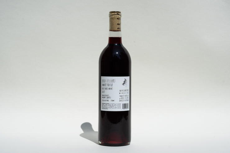 A bottle of 2023 Amore Rosso by Broc Cellars, a refined Italian red wine, is set vertically against a light plain backdrop. The Barbera Dolcetto blend includes a cork and a label with black text and a small logo. Shadows fall to the left on the surface.