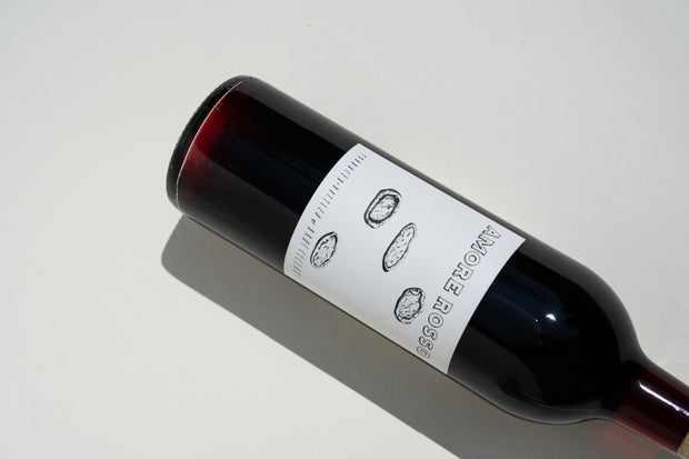 A bottle of 2023 Amore Rosso by Broc Cellars lies sideways on a light background. The label features line drawings of three corks. This dark red Barbera Dolcetto blend is lit from above, casting a shadow.