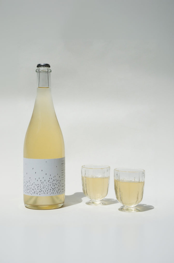 A tall clear bottle of Love Sparkling by Broc Cellars stands beside two Broc wine glasses, all filled with the same light yellow liquid. The bottle has a minimalist white label adorned with small black dots against a softly lit background.
