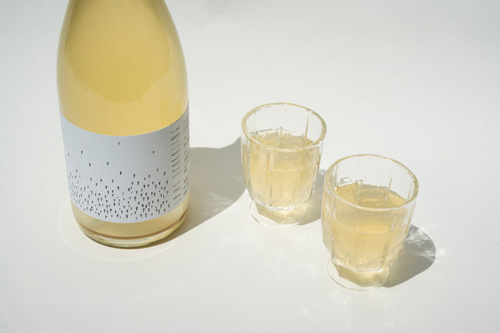 A bottle of pale yellow Love Sparkling from Broc Cellars with a minimalist gray label featuring small black dots sits beside two wine glasses on a white surface, both glasses containing the same inviting yellow liquid.