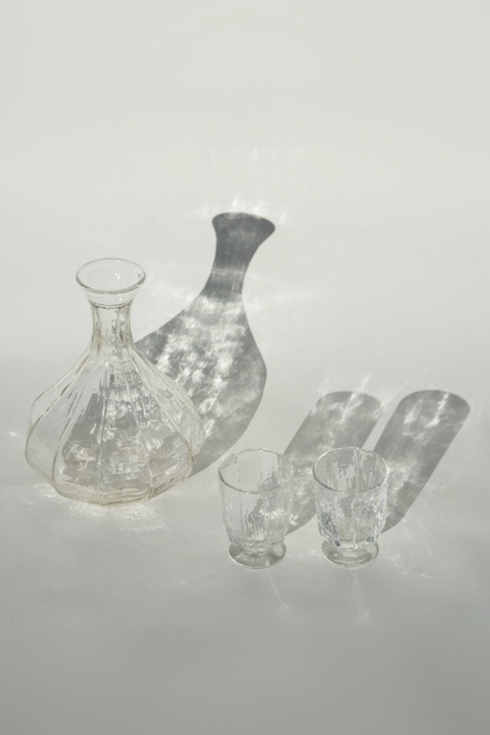 The Broc Cellars Decanter + Glass Set, featuring a clear handcrafted glass decanter and two casual wine glasses, rests on a light surface with elongated shadows and visible light reflections.