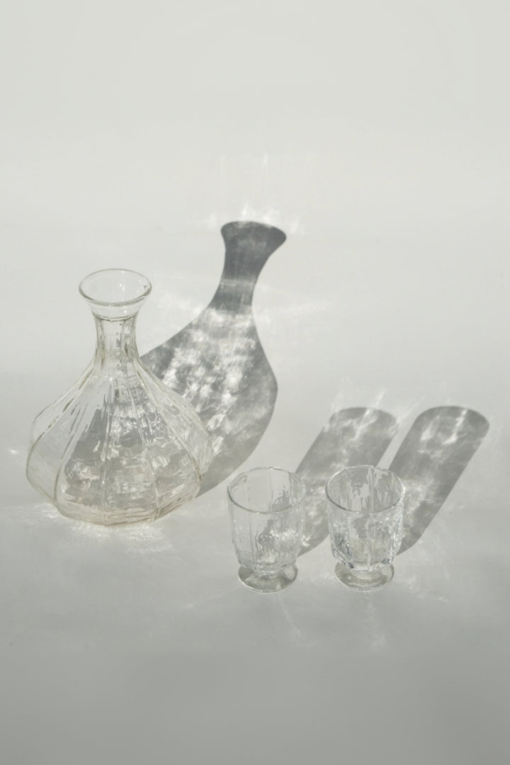 The Broc Cellars Decanter + Glass Set, featuring a clear handcrafted glass decanter and two casual wine glasses, rests on a light surface with elongated shadows and visible light reflections.