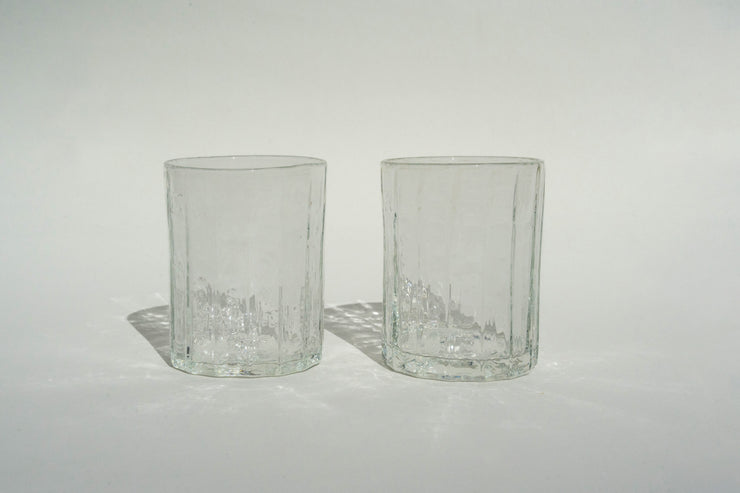 Two clear Manhattan Glass Set tumblers from Broc Cellars sit side by side on a light surface, casting subtle shadows under overhead lighting. The neutral background accentuates the transparency and texture of the handblown glass.