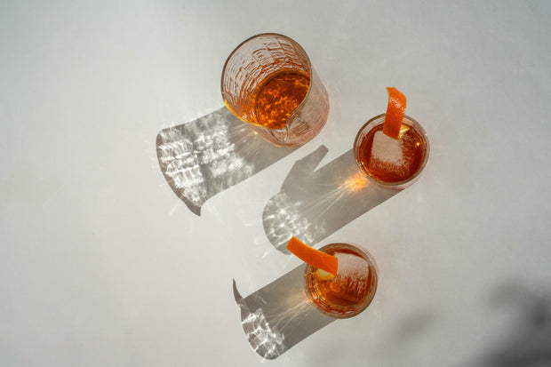 A handblown glass pitcher and two cocktail glasses, part of the Manhattan Glass Set by Broc Cellars, filled with amber liquid rest on a white surface. Each glass has an orange peel garnish, while the light casts long shadows, creating patterns that dance elegantly across the set.