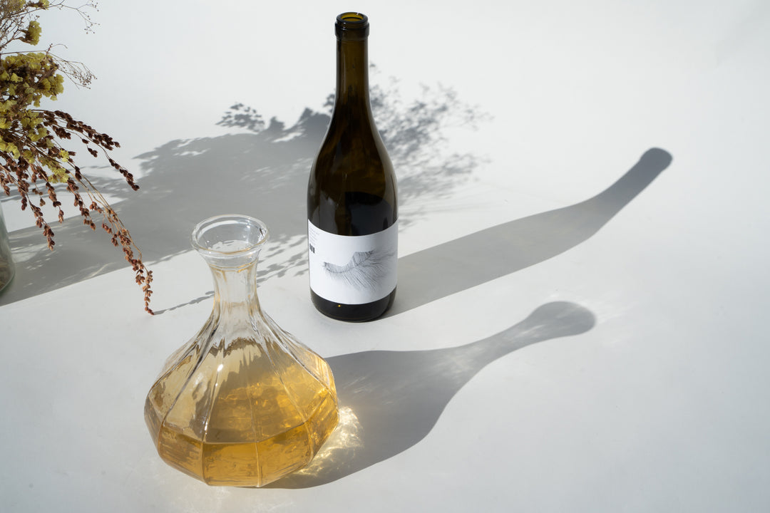 A half-filled glass decanter with a light amber liquid, resembling a California Chardonnay, is in the foreground. Behind it stands a dark wine bottle with a white label from Broc Cellars 2023 Michael Mara Chardonnay. Shadows dance on the surface while dried flowers peek from the left.