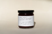 A jar with a black lid features handwritten text. The label highlights organic ingredients: figs, sugar, Broc Angelica Wine, ginger, lemon, and lemon thyme. It prominently displays June Taylor Mission Fig + Angelica Jam by Broc Cellars with an Oakland, CA address and website.