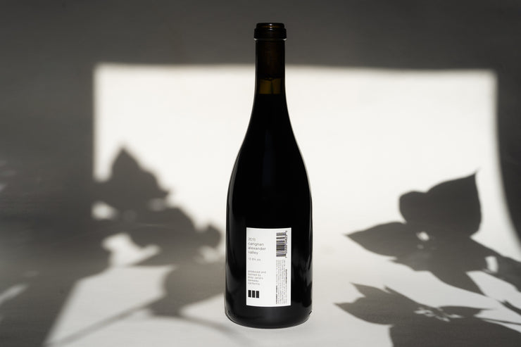 A dark wine bottle of Broc Cellars 2015 Old Vine Carignan sits on a white background, casting leaf-like shadows. The back label with text and barcode is visible, and the lighting highlights its whole cluster fermentation technique for Carignan plants.