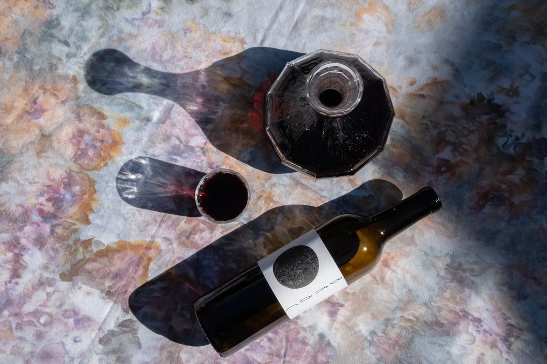 A half-full decanter, wine bottle, and glass of 2022 Broc Massa Cabernet Sauvignon sit on a floral tablecloth, casting shadows. The bottle from Broc Cellars features a minimalist label with a black circle design, originating from the Massa Vineyard in California.