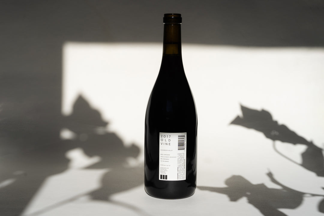 A dark wine bottle of Broc Cellars’ 2018 OLD VINE CARIGNAN stands on a white surface against a white background. The back label reads 2018 Old Vine, alluding to the storied heritage of ancient vines from which it was crafted. Shadows of leaves and branches are softly cast across the scene.