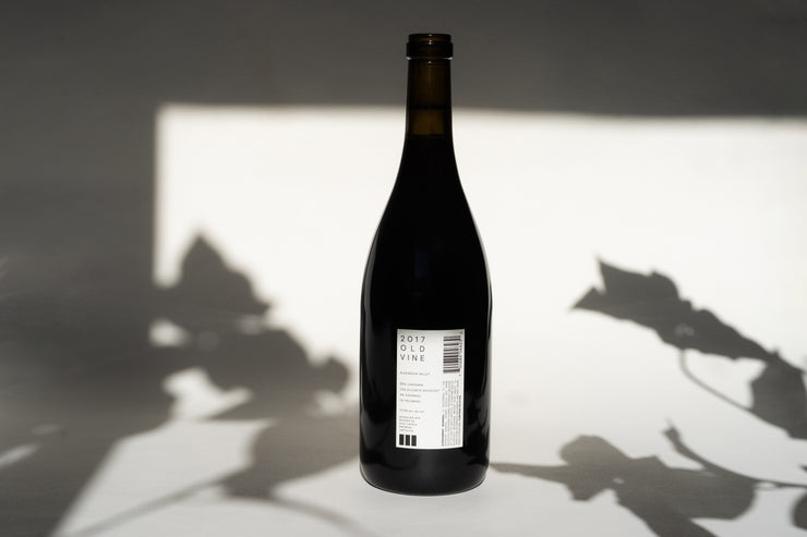 A bottle of 2017 OLD VINE CARIGNAN by Broc Cellars, crafted with native yeasts, sits on a flat surface. Shadows of leaves add texture to the background. The label and barcode face forward, reflecting its ancient vineyard origin.