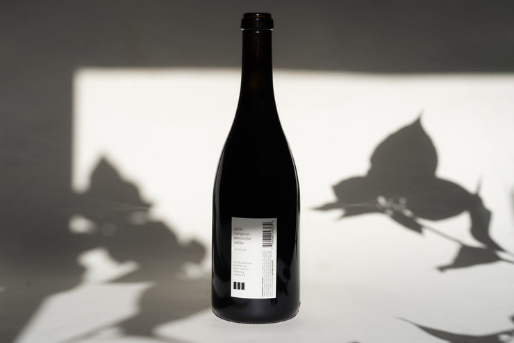 A dark glass wine bottle labeled 2013 OLD VINE CARIGNAN by Broc Cellars stands upright amid leaf shadows, reminiscent of an ancient vineyard, with text and a barcode on the white label.