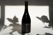 A bottle of 2010 LUNA MATTA MOURVEDRE from Broc Cellars, with a dark floral label, stands against leafy shadows. Its bold presence evokes Paso Robles terroir, with dramatic lighting highlighting the rich character as it casts its distinct shadow.