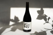 The 2018 NERO D’ AVOLA from Broc Cellars features a dark wine bottle with a black and white drawing of a badger on the label. Against a light background, leaf shadows subtly dance, reflecting its earthy Mendocino origins.