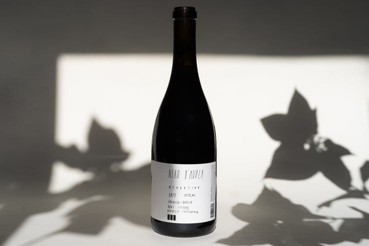 A 2015 Nero d’Avola bottle from Broc Cellars stands upright, partially lit, with a monochrome label hinting at its origins. Leaf shadows add texture and contrast to the scene.