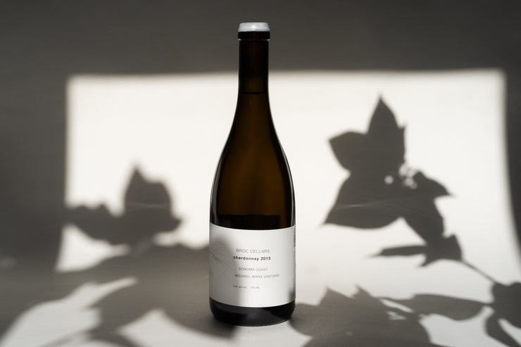 A 2015 Michael Mara Chardonnay from Broc Cellars with a white label sits on a surface. The text is partly legible as shadows from leaves play decoratively in the background. Soft lighting accentuates the glass.