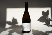 A tall, slender bottle of 2017 MICHAEL MARA CHARDONNAY by Broc Cellars features a minimalist white label, set against a light backdrop. Shadowy leaf patterns on the surface create an artistic contrast.