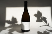 A wine bottle labeled 2014 Michael Mara Chardonnay from Broc Cellars sits on a surface, casting a shadow on the wall, with leaf shadows dancing in the background.