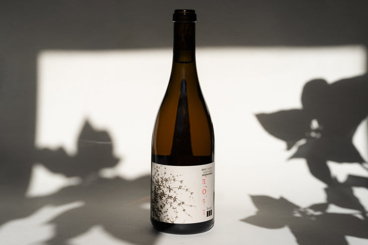 The 2013 CASSIA GRENACHE ROSE by Broc Cellars stands on a surface with its minimalist floral label, featuring ROSÉ, capturing the essence of McDowell Valley. Soft leaf shadows dance on the light background, adding mystery.