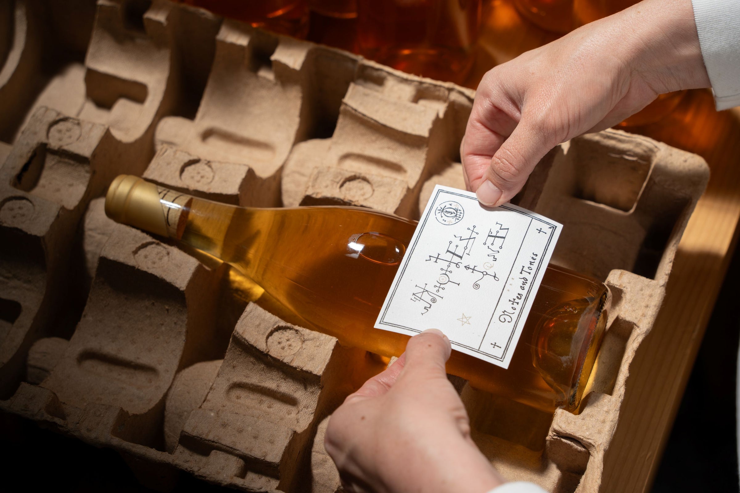 A person places a label on a bottle of golden-colored liquid, possibly wine or cider, inside a molded cardboard shipping container with other similar bottles visible in the background.