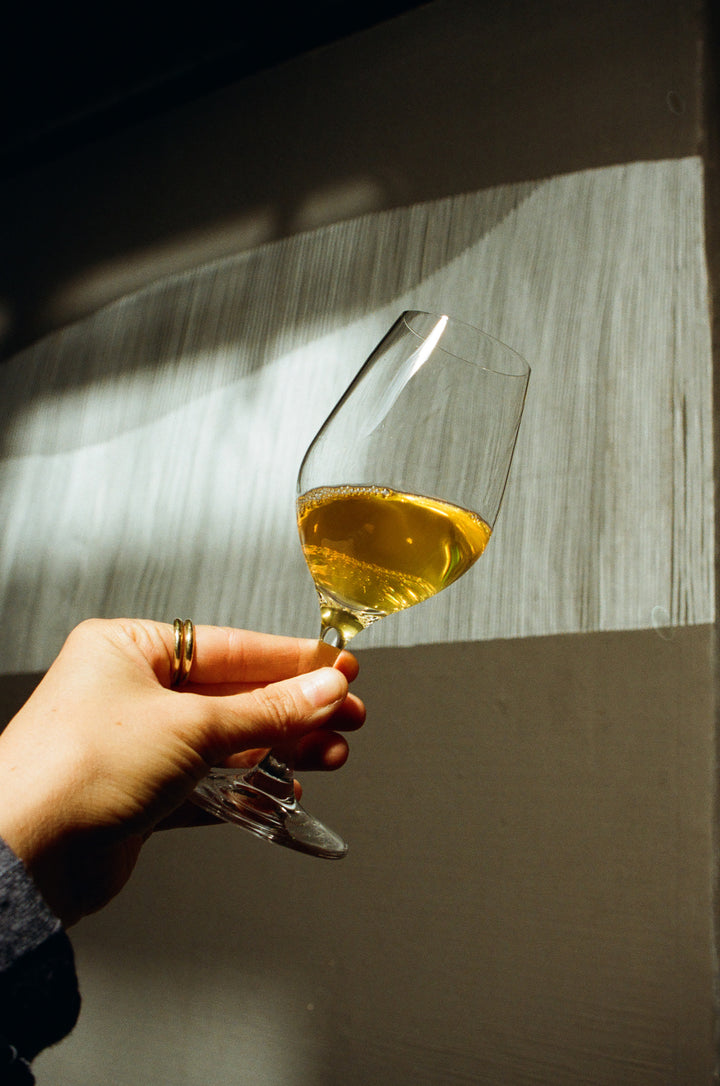 A hand adorned with two gold rings gracefully holds a wine glass filled with golden liquid, possibly Notes &amp; Tones V.2 from Broc Cellars. The textured wall casts intriguing patterns of light and shadow, enhancing the elegance of the scene.