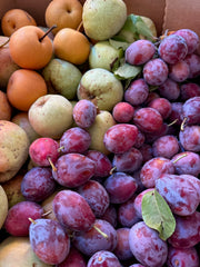 A close-up of a box filled with fresh fruit, including green pears and small brown Asian pears, plus reddish-purple plums ideal for June Taylor x Fox Hill Santa Rosa Plum w/ Lavender from Broc Cellars. Some fruits have leaves attached.