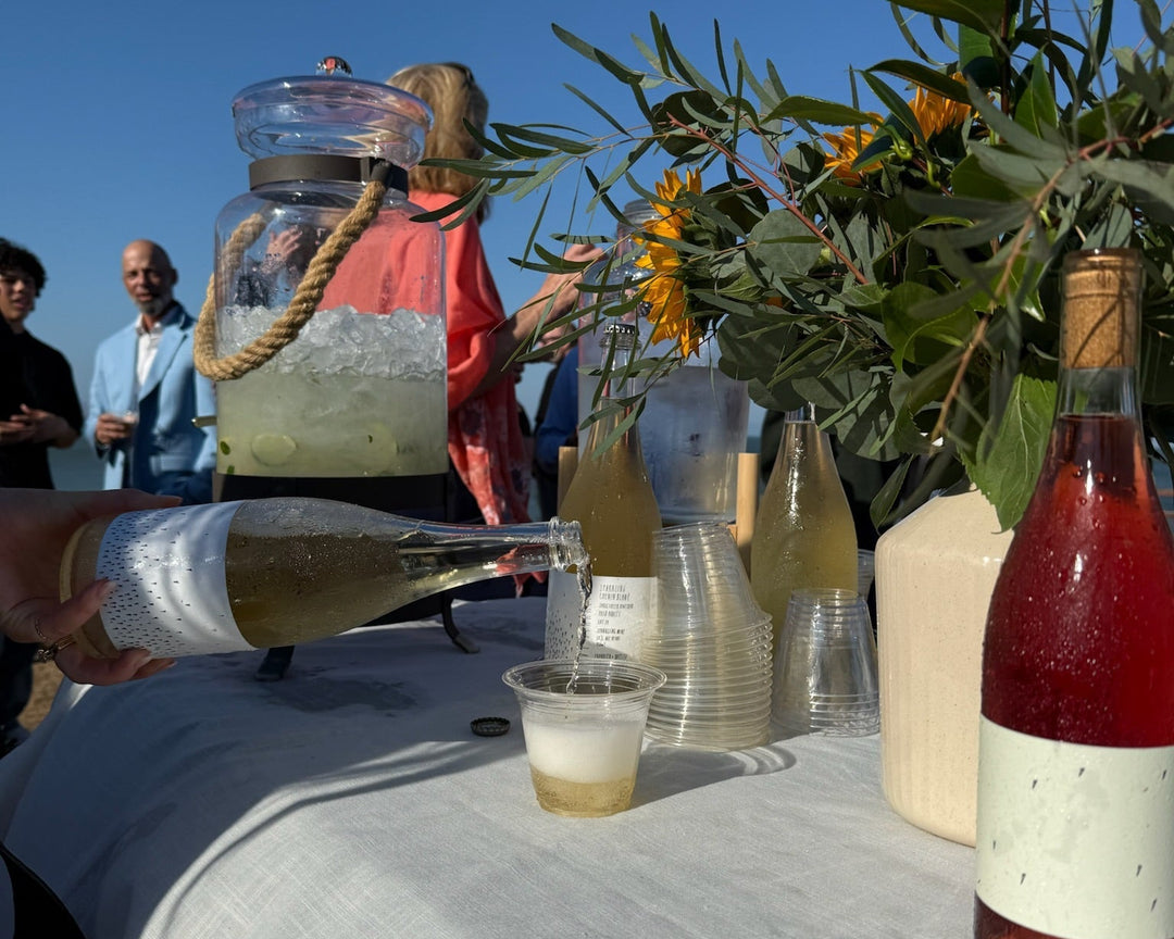 A hand pours white wine into a plastic cup on a table with a beige tablecloth, glass bottles, cups, a vase with flowers, and a large drink dispenser. Several people stand in the background outside under a clear blue sky.