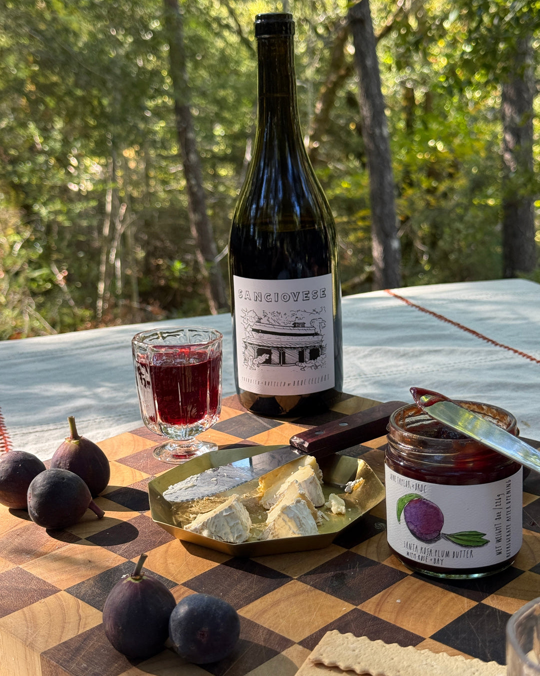 A bottle of Sangiovese wine, a glass of red wine, figs, a wedge of brie cheese with a knife, fig jam, and crackers are arranged on a wooden board outdoors with trees in the background.