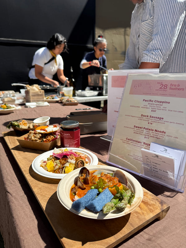 A wooden board with assorted plated dishes is displayed on a table outdoors. A menu stands upright near the dishes. Two people prepare food in the background under bright sunlight.