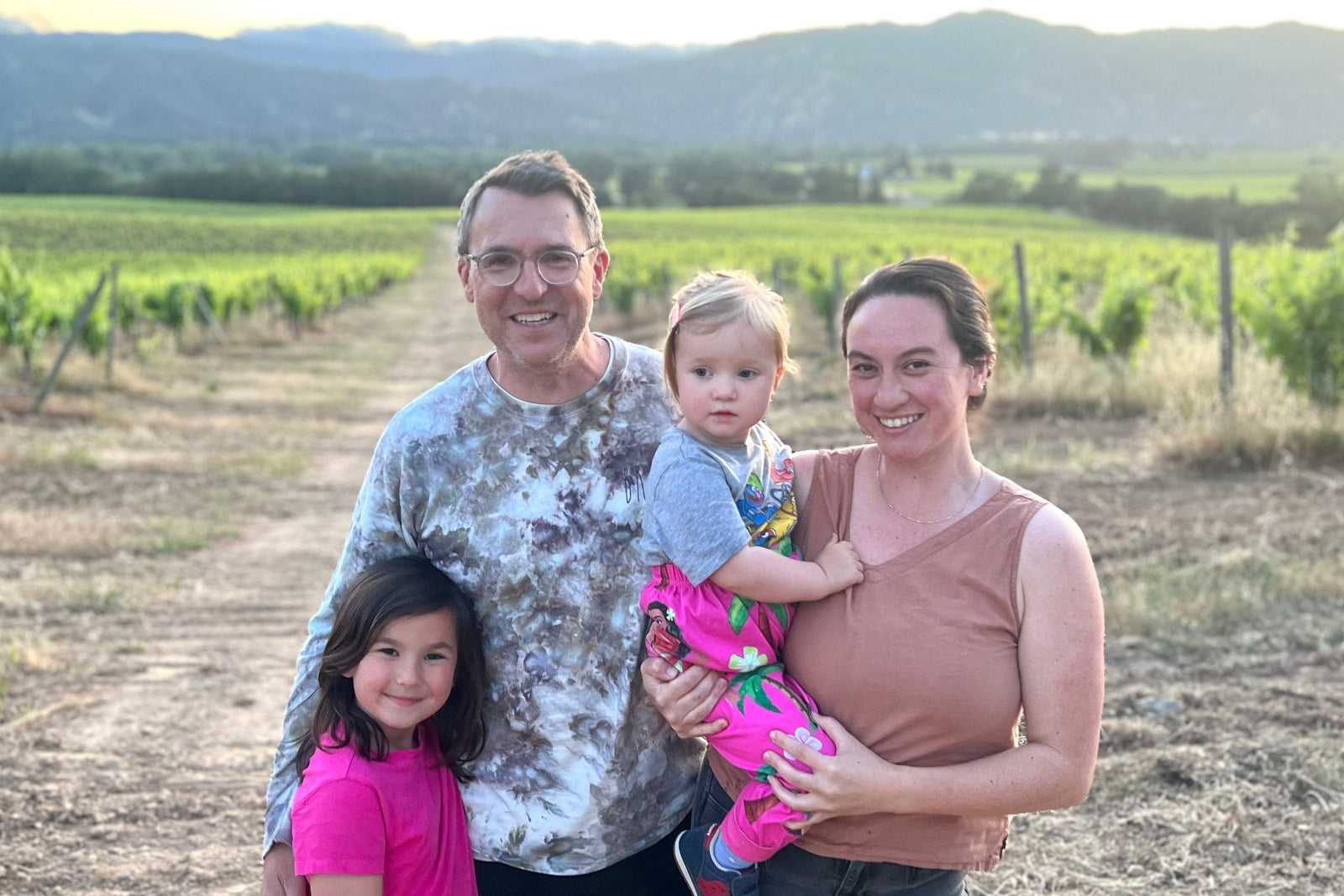 A family of four stands on a dirt path in a vineyard. A man and woman hold a toddler, with an older child standing beside them. The background features rows of grapevines and distant hills under a partly cloudy sky.