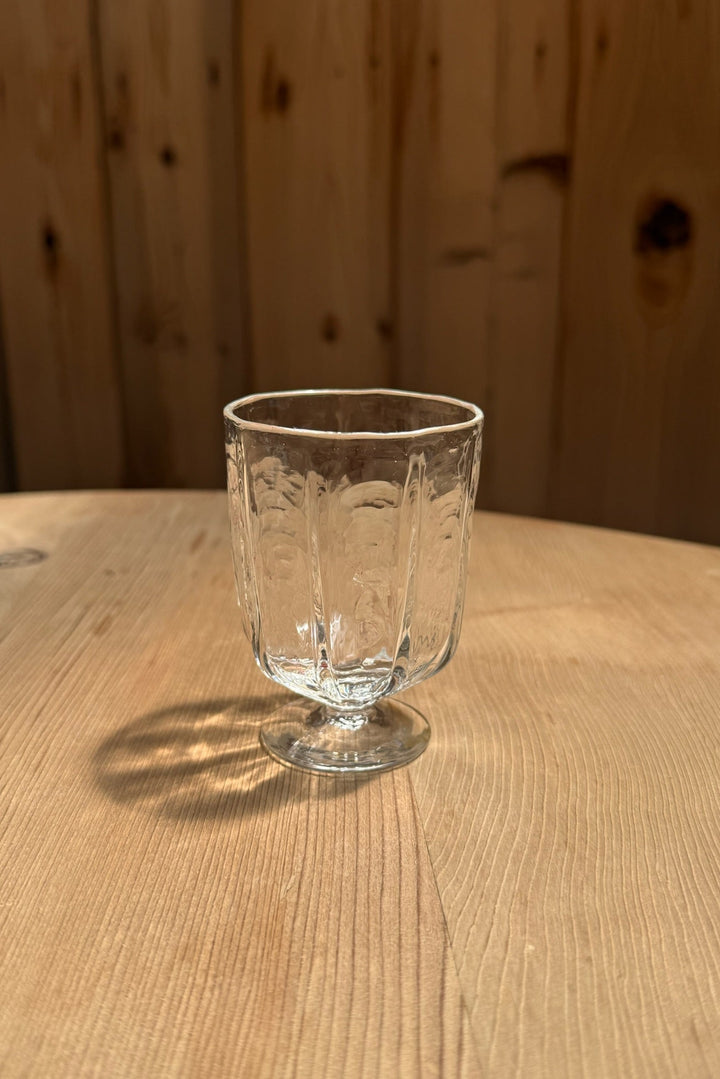 A Broc Glass by Broc Cellars, featuring an embossed pattern, sits on a light wooden table with sunlight casting its handblown form&