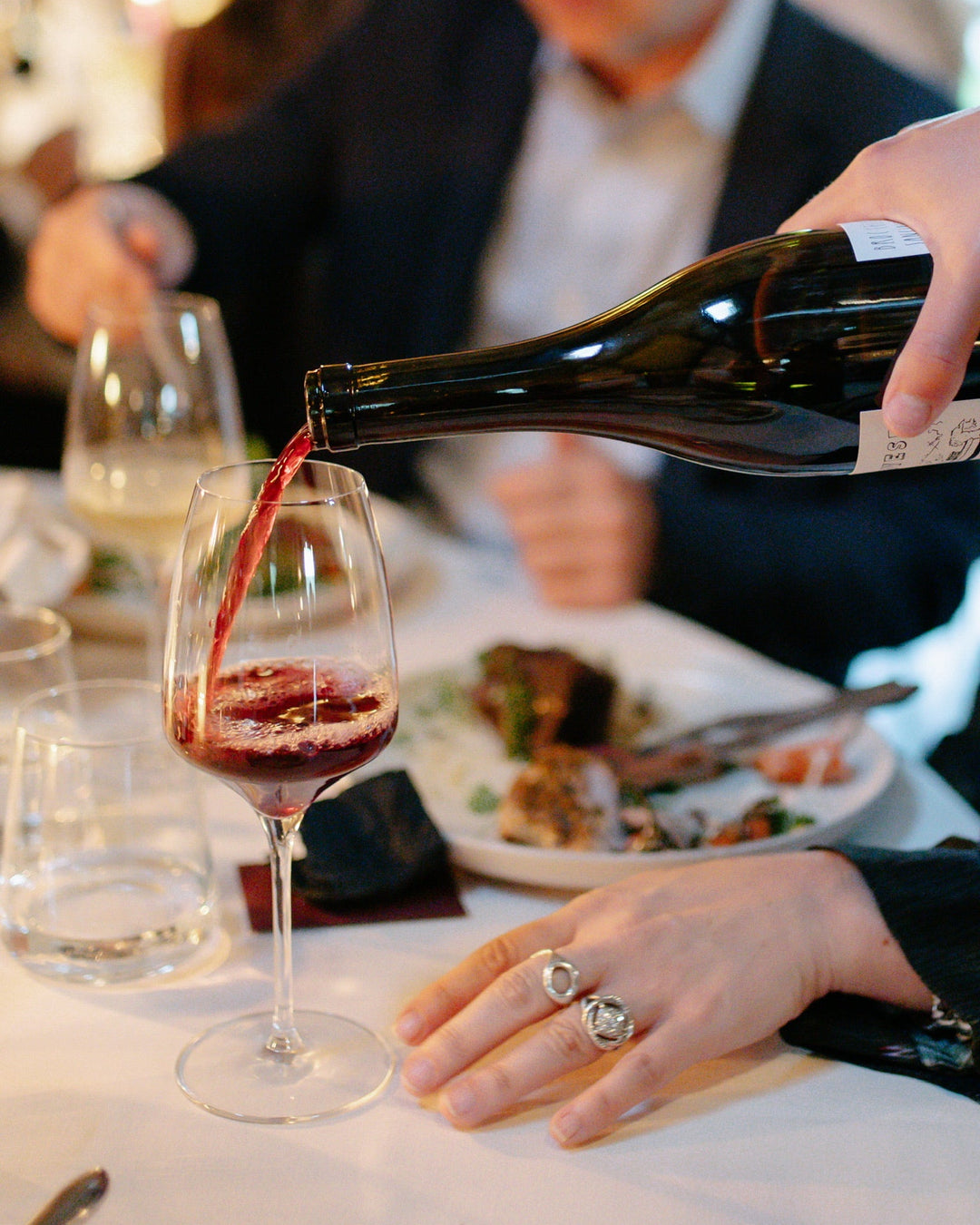 A hand pours red wine from a bottle into a wine glass on a set dining table with glasses, plates of food, and people seated, one person’s hand resting near the glass.