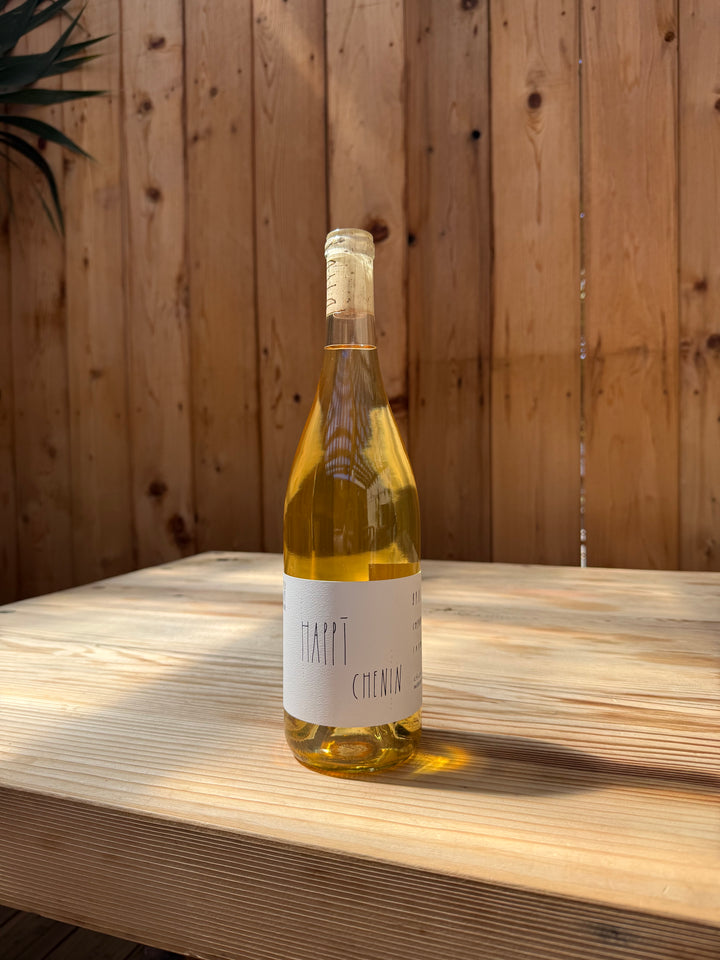 A bottle of Broc Cellars’ 2021 Happī Chenin rests on a wooden table, sunlight streaming in and a wooden fence in the background, evoking the serene charm of Massa Vineyard.