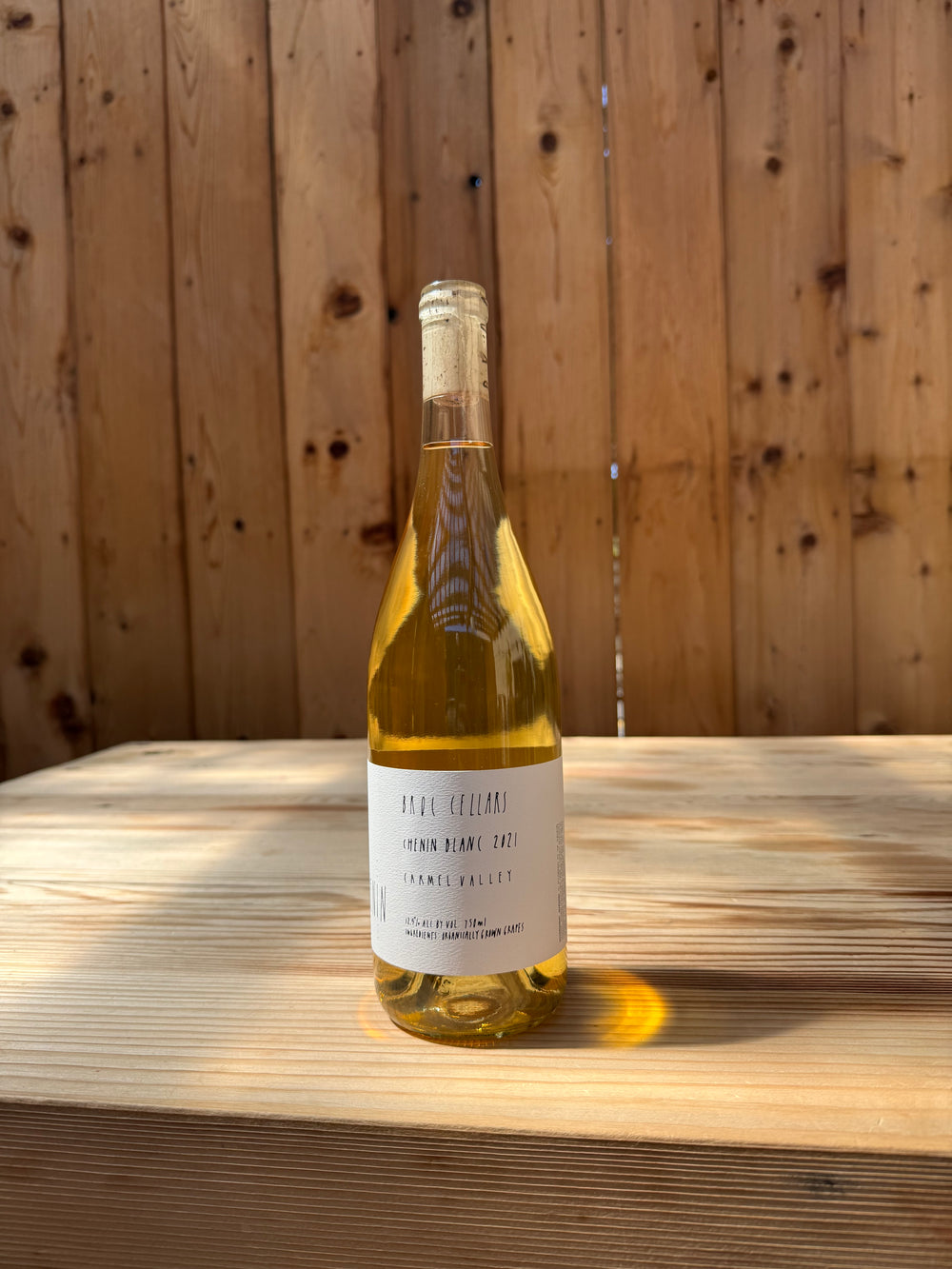 A bottle of white wine—2021 Happī Chenin from Broc Cellars—stands on a wooden surface with a wooden fence in the background. Sunlight illuminates the bottle, casting a yellow reflection on the wood. The label is partially visible but not entirely readable.