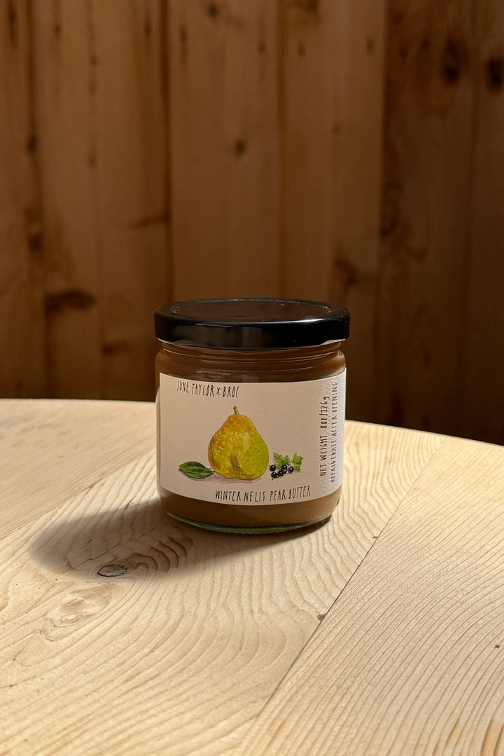 A glass jar with a black lid labeled "June Taylor x Fox Hill Winter Nelis Pear Butter" by Broc Cellars sits on a light wooden surface, its label highlighting this low-sugar fruit preserve with an illustration of a pear.