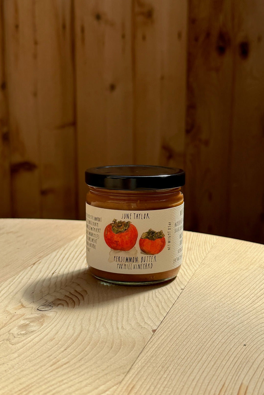 A glass jar of Broc Cellars June Taylor Persimmon Butter sits on a light wooden table. The label shows persimmon illustrations and text, with an out-of-focus wood paneling background.