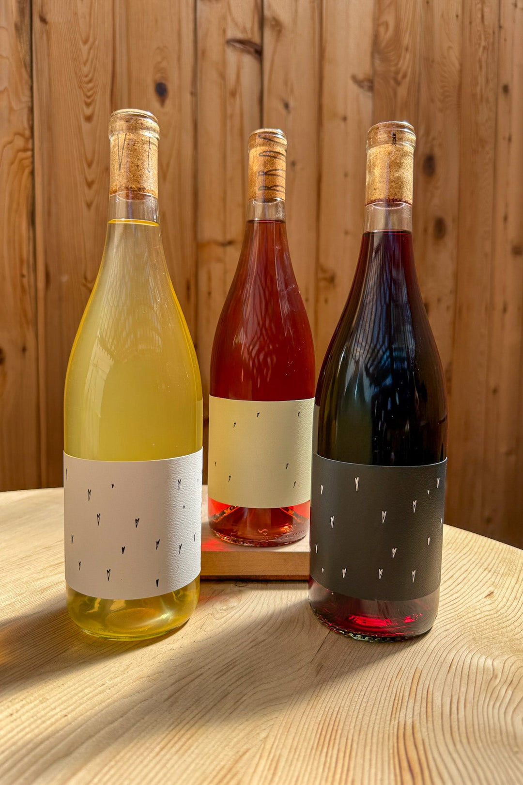 Three Broc Cellars Love Trio wine bottles—white, rosé, and red—each with minimalist black-mark labels, sit on a wooden table. Perfect as a gift box or party wine. A wooden wall is in the background.