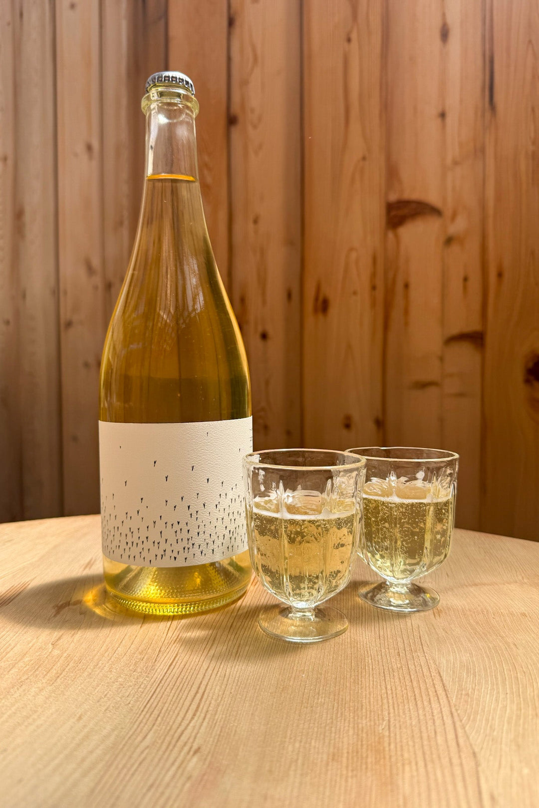 A bottle of Broc Cellars’ Love Sparkling with Wine Glasses sits on a light wooden table beside two filled glasses, set against a wooden panel wall—a stylish scene for celebrating in 2023.